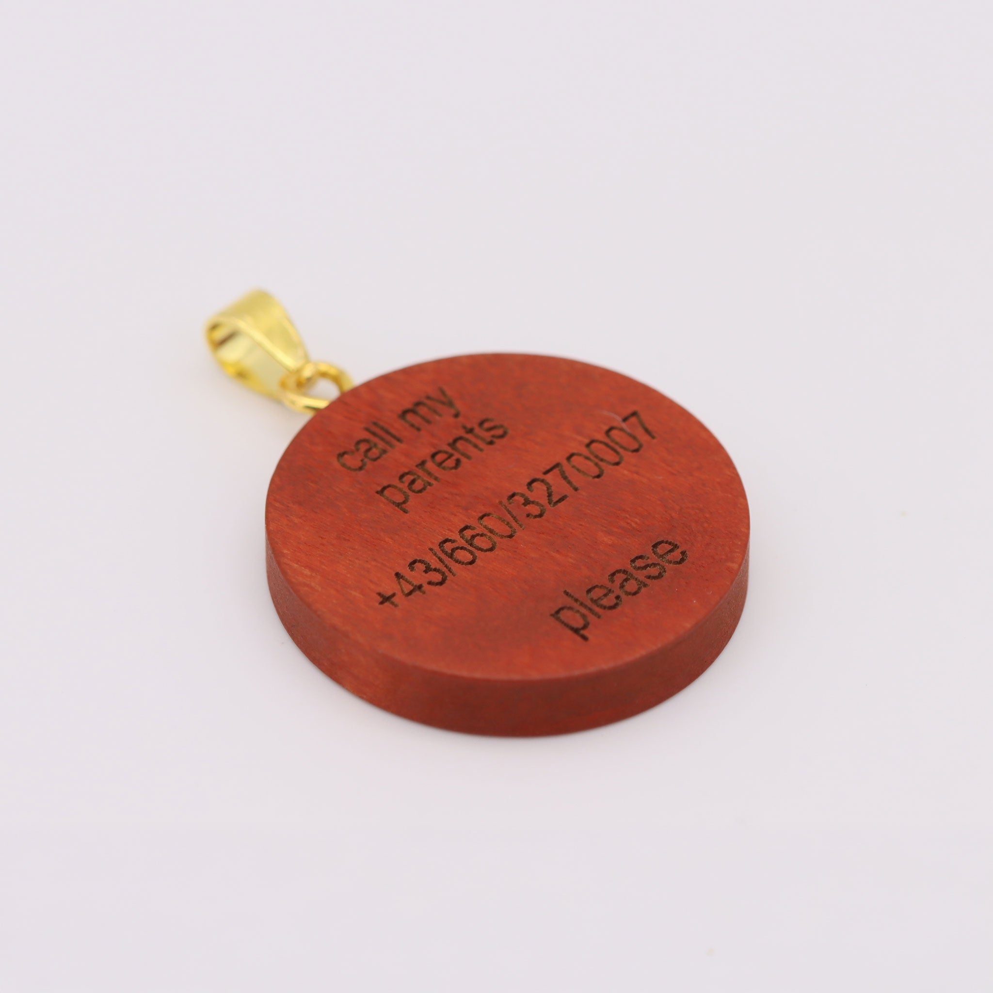 Eine Nahaufnahme der gravierten Holzrückseite einer Hundemarke. Sie zeigt die klare, lasergravierte Aufschrift 'call my parents', eine österreichische Telefonnummer (+43/660/3270007) und 'please'. Die Marke ist an einem goldenen Karabinerverschluss befestigt