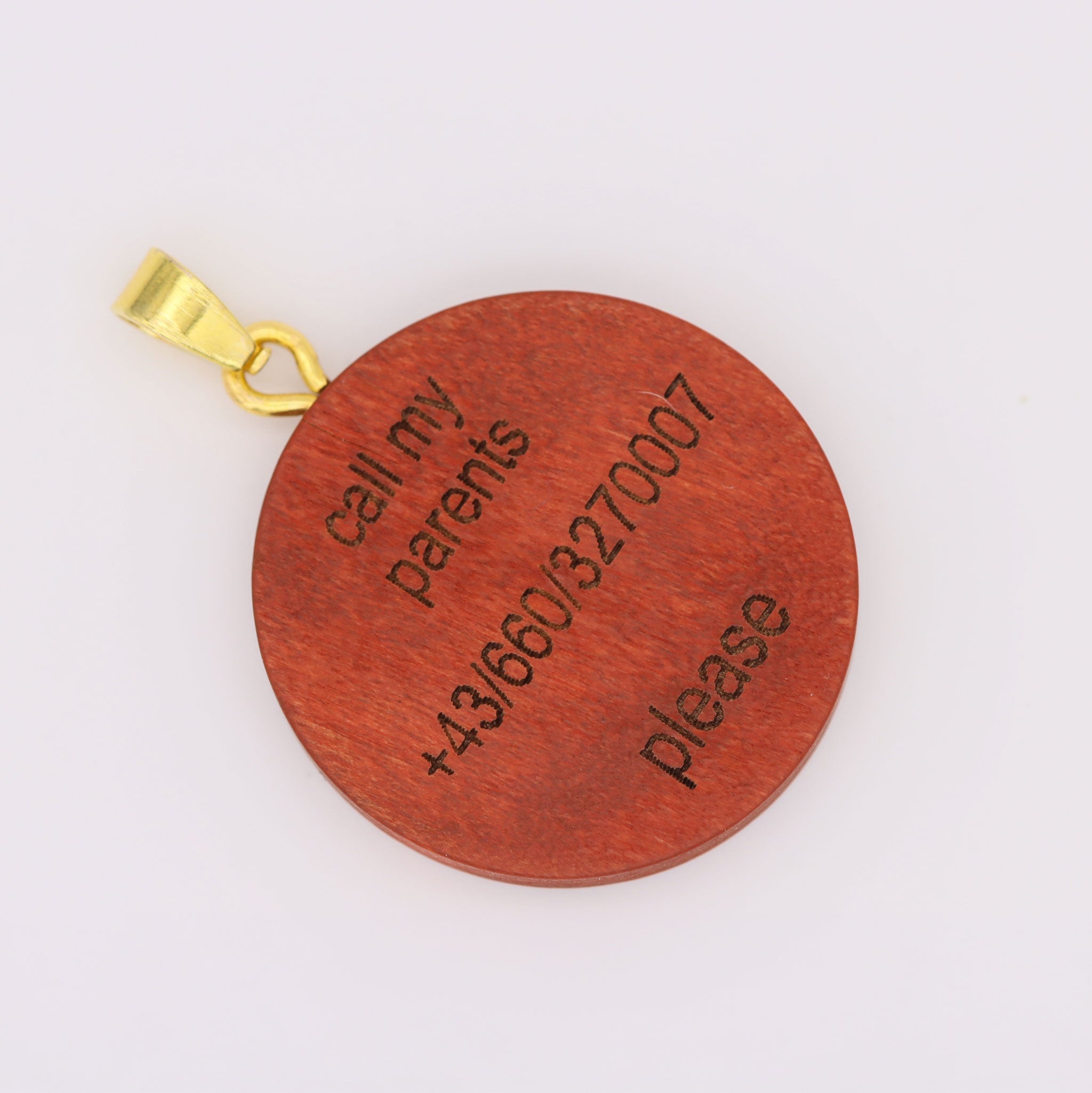 Eine Nahaufnahme der gravierten Holzrückseite einer Hundemarke. Sie zeigt die klare, lasergravierte Aufschrift 'call my parents', eine österreichische Telefonnummer (+43/660/3270007) und 'please'. Die Marke ist an einem goldenen Karabinerverschluss befestigt