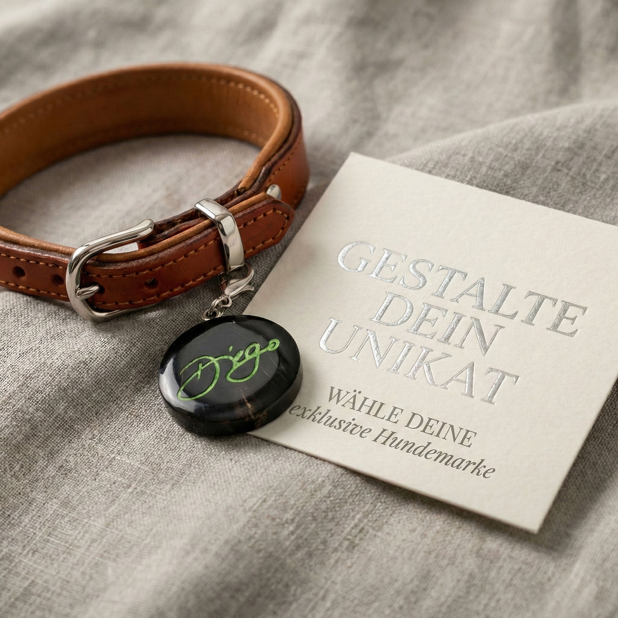 Individuelle Hundemarke aus schwarzem Büffelhorn mit grüner Namensgravur an einem braunen Lederhalsband mit Silber-Beschlägen, präsentiert auf edlem Leinenstoff neben einer Karte mit der Aufschrift „Gestalte dein Unikat“.