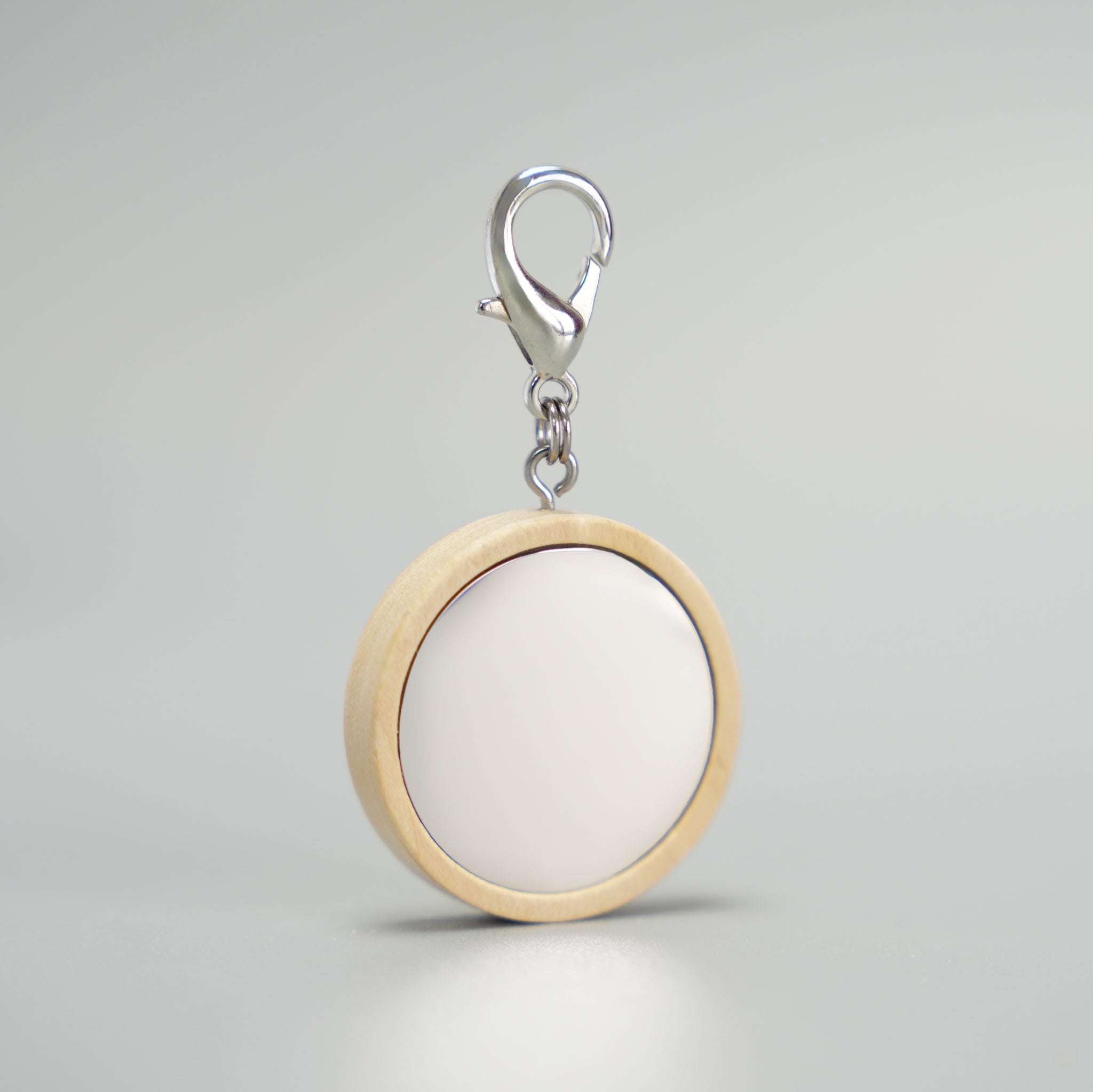 Runde, blanko Hundemarke mit hellem Holzrahmen, mattem Silberinlay und silbernem Karabinerverschluss vor einem grauen Hintergrund. Bereit für die Gravur eines Hundenamens und Kontaktdaten.