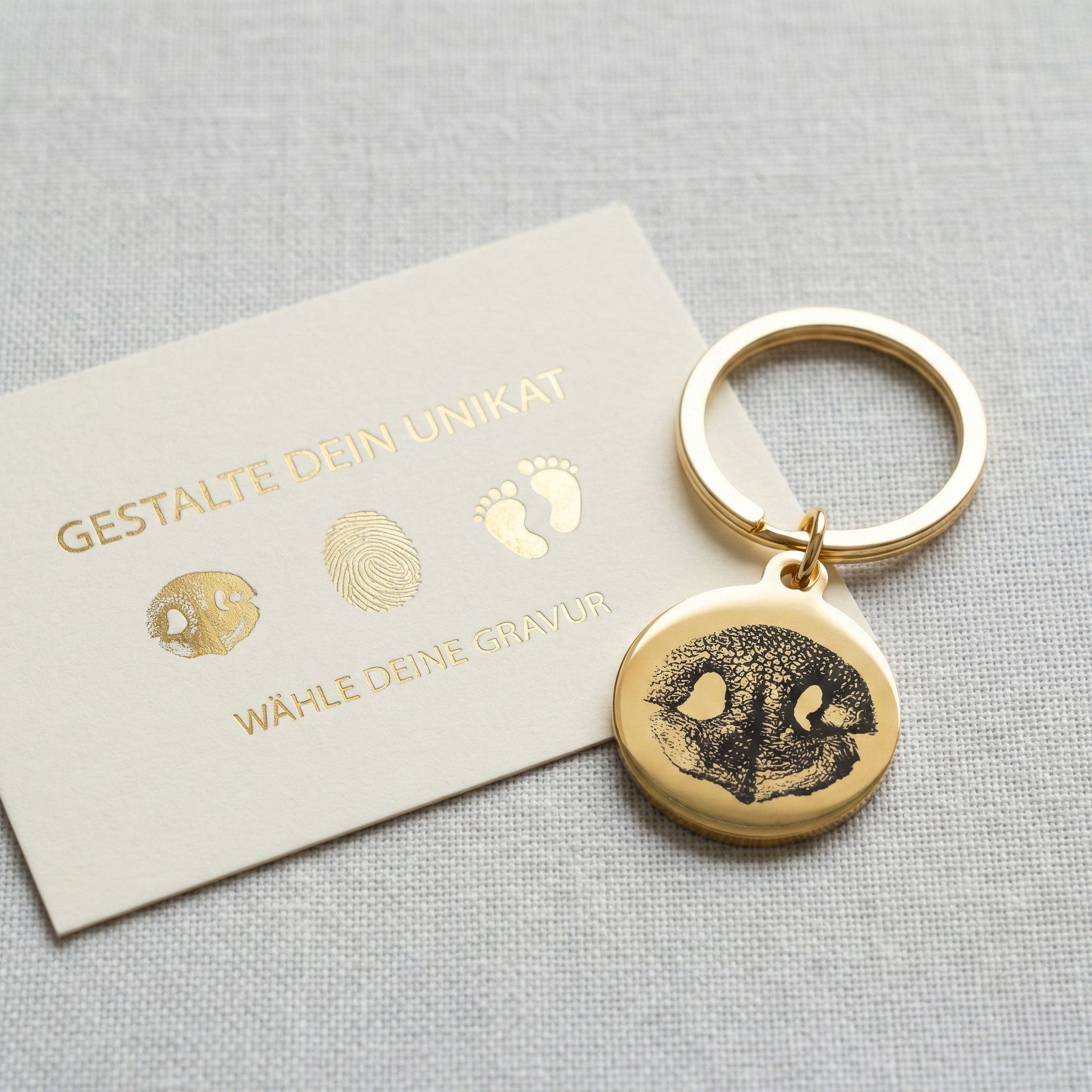eine boutique-design karte die die Auswahl an Gravurmöglichkeiten für den rechts liegenden Schlüsselanhänger zeigt. auf dem goldenen, runden Schlüsselanhänger ist der Nasenabdruck eines Hundes graviert