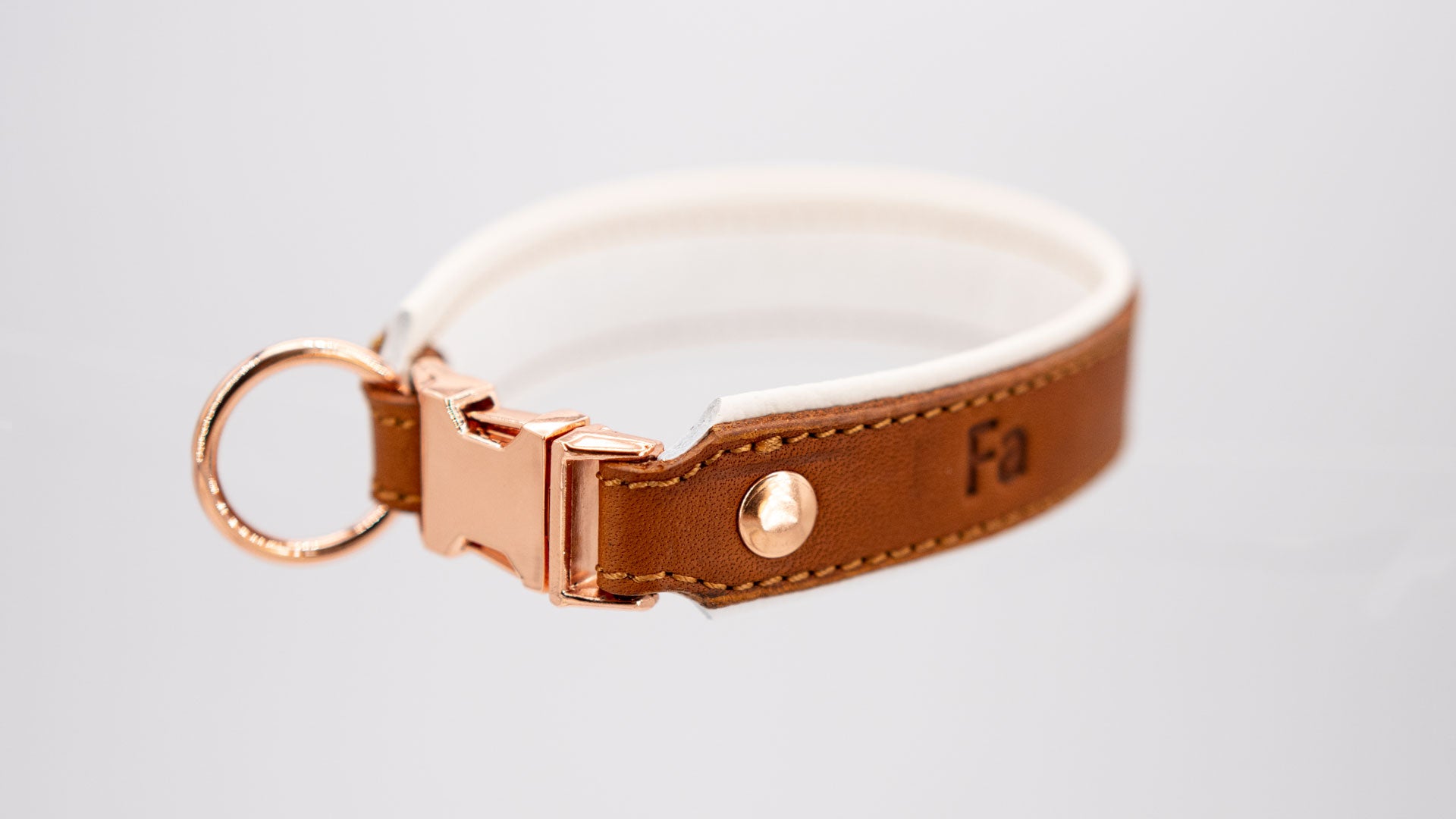 handgefertigtes fettlederhalsband in cognac mit weiß hinterlegt, seitlich gedreht mit rose goldenem steckverschluss. man kann 2 geprägte buchstaben erkennen