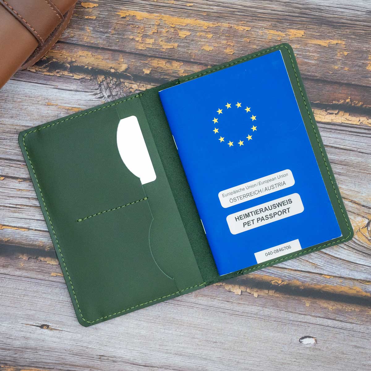 Geöffnete Leder-Impfpasshülle in Grün, rechts befüllt mit einem blauen EU-Heimtierausweis und Sicht auf die präzise, aufwendige Kantenverarbeitung.