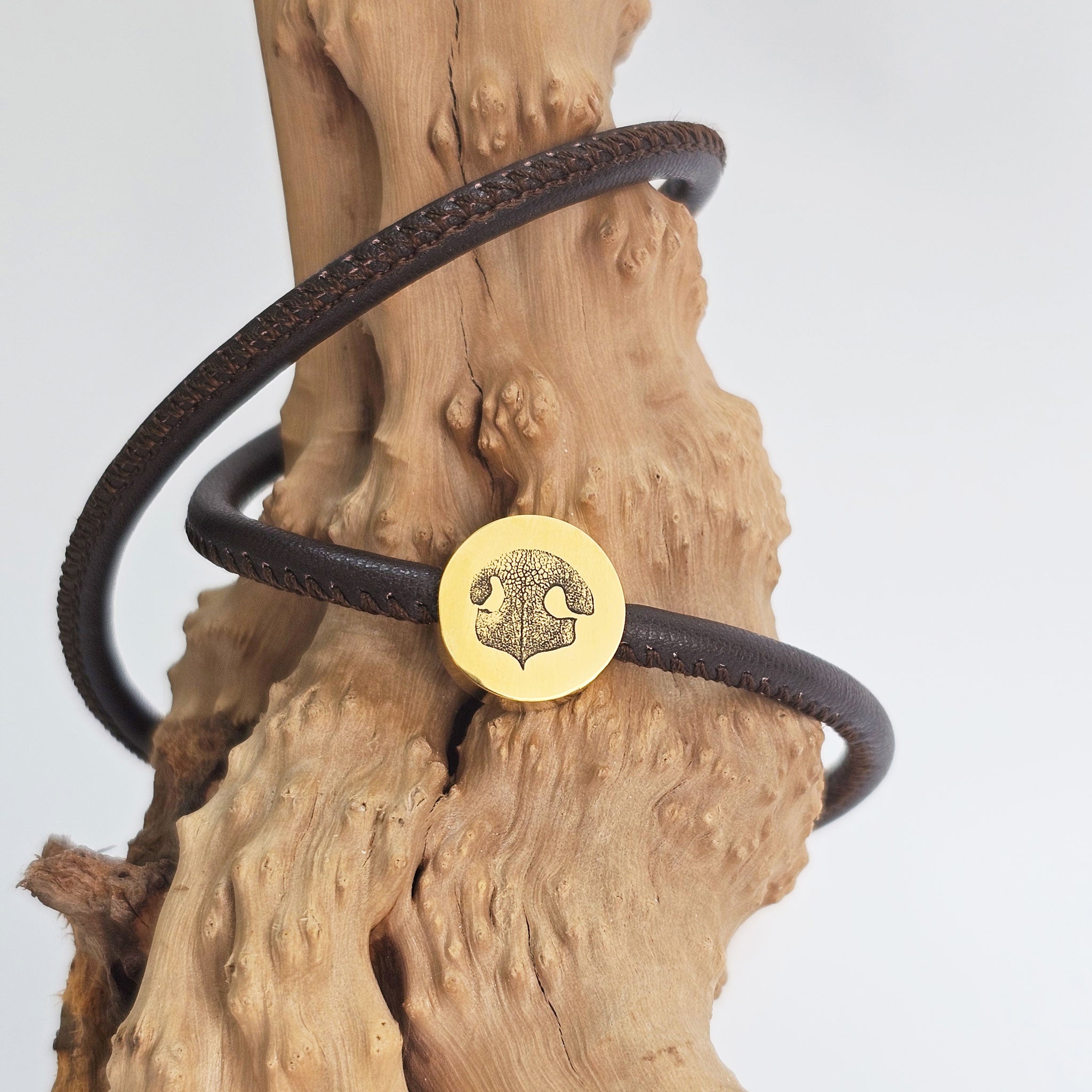 Dunkelbraunes Wickelarmband aus Leder mit goldener Hundenasen-Gravur, dekoriert auf einem Stück Treibholz