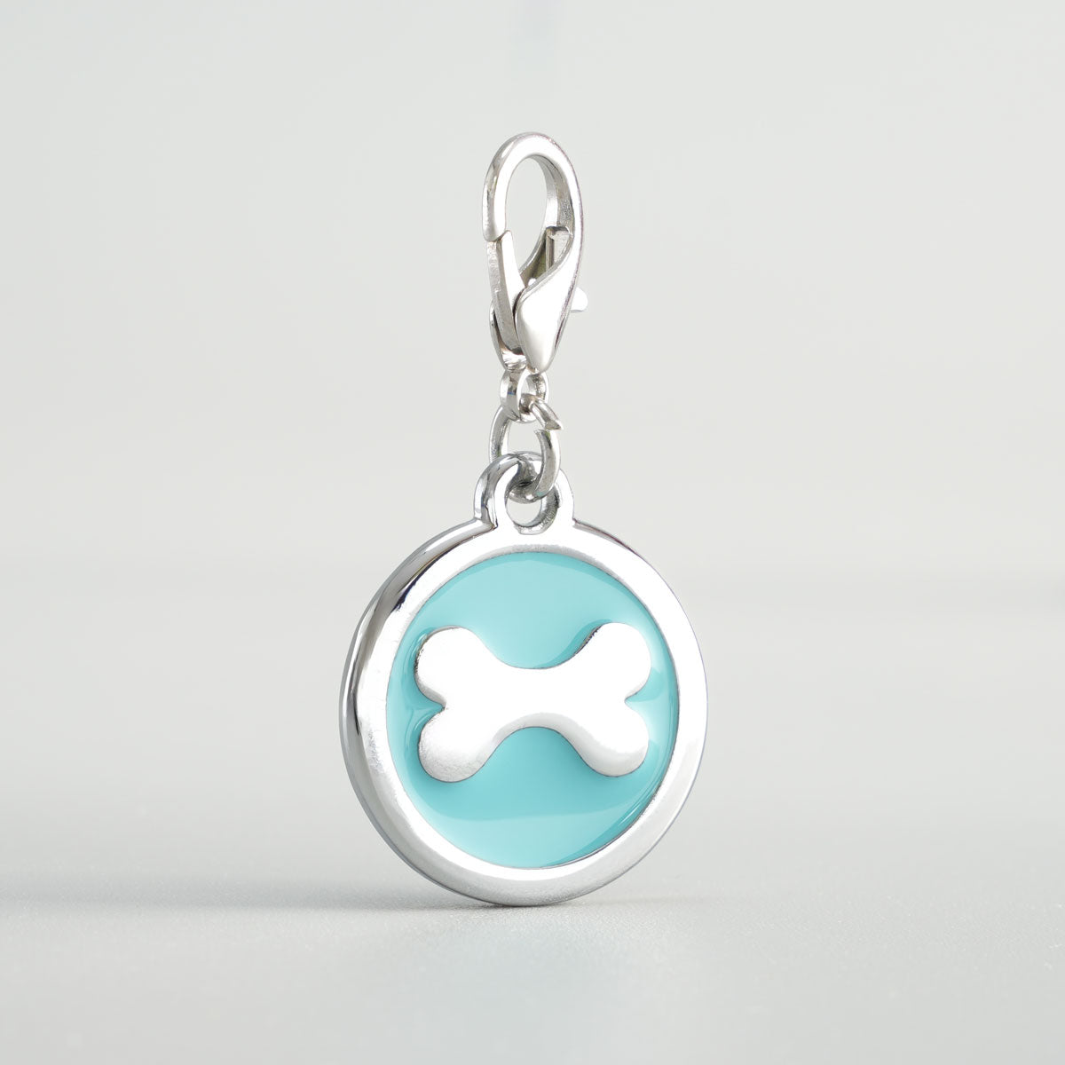 Detailansicht einer hellbaluen, runden Hundemarke aus verchromtem Druckguss mit silbernem Knochen-Inlay und Karabiner