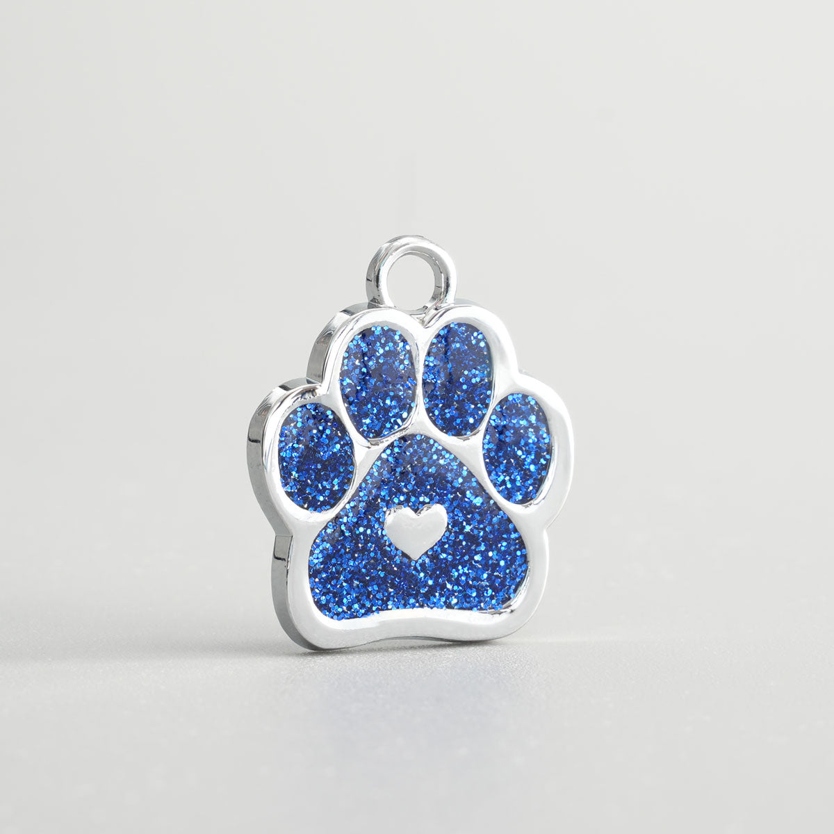 Nahaufnahme einer verchromten Hundemarke in Pfotenform mit blauem Glitzer-Inlay und silbernem Herz in der Mitte.