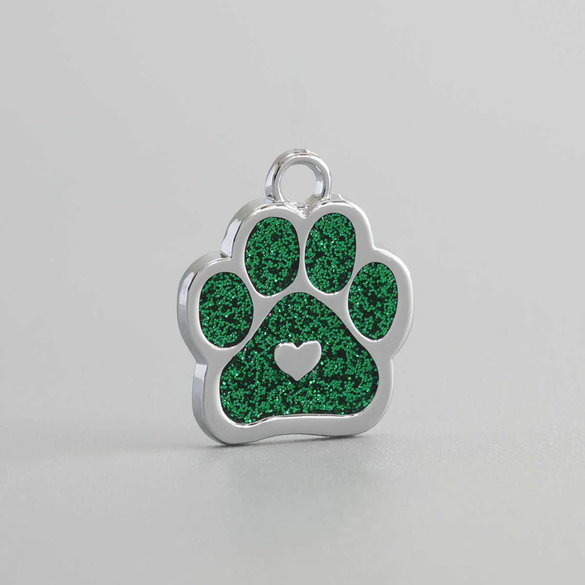 Nahaufnahme einer verchromten Hundemarke in Pfotenform mit grünem Glitzer-Inlay und silbernem Herz in der Mitte.