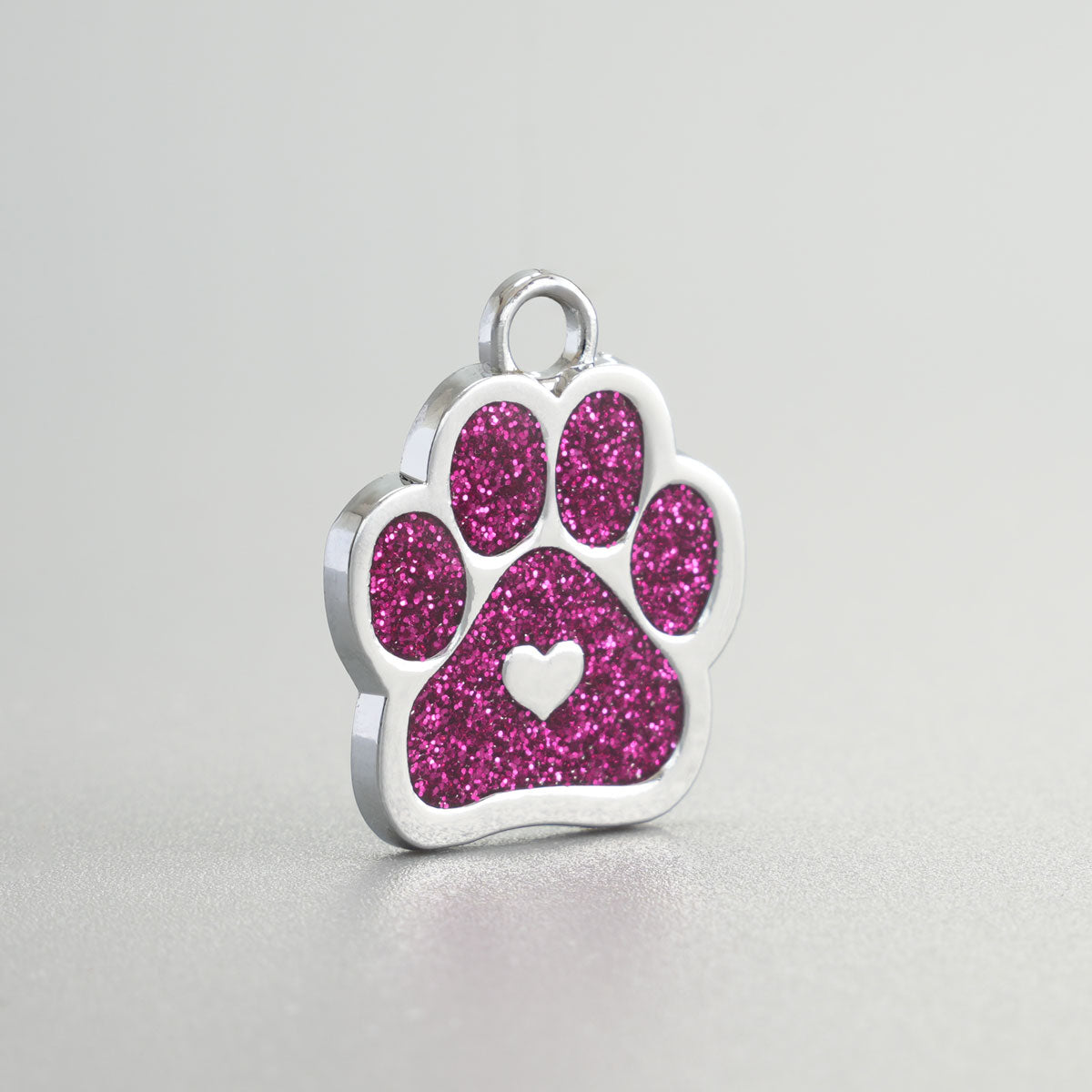 Nahaufnahme einer verchromten Hundemarke in Pfotenform mit magenta Glitzer-Inlay und silbernem Herz in der Mitte.