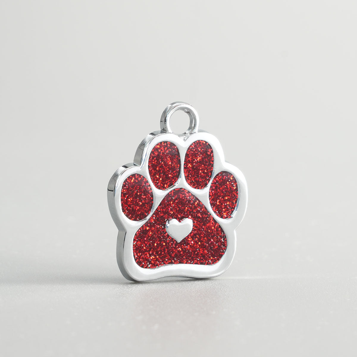 Nahaufnahme einer verchromten Hundemarke in Pfotenform mit rotem Glitzer-Inlay und silbernem Herz in der Mitte.