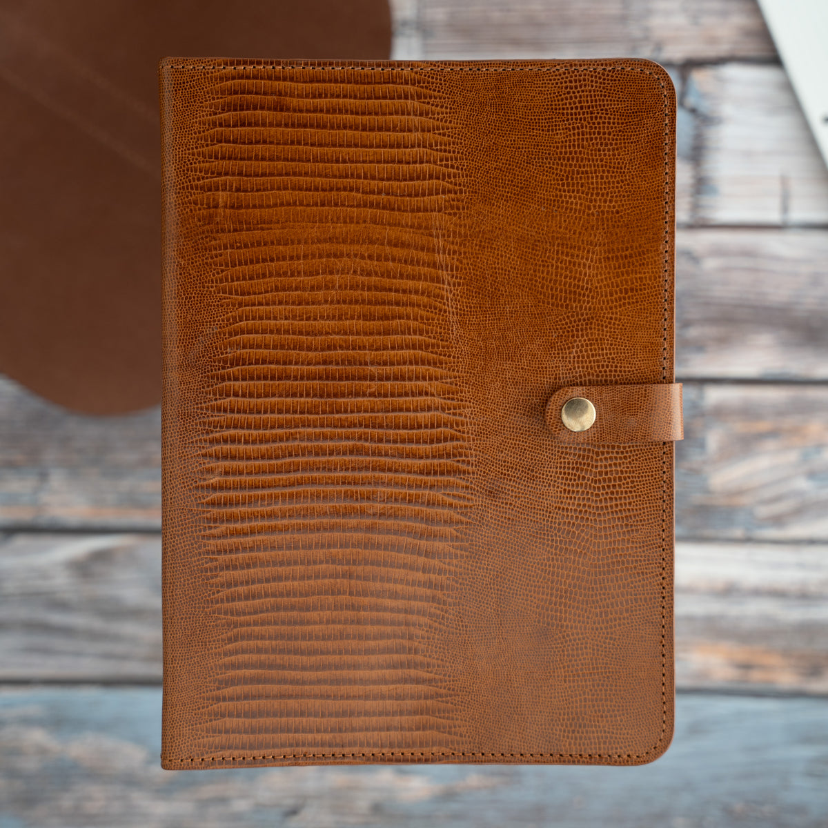 Geschlossenes A5-Leder-Ringbuch aus cognacfarbenem Kalbsleder mit Echsenprägung und Lasche mit Messing-Druckknopf.