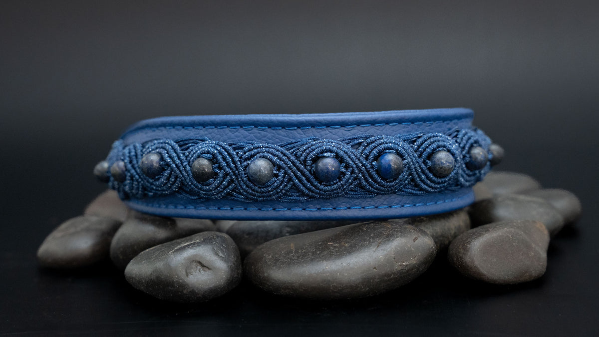 Gesamtansicht im Querformat: blaues Nappaleder-Halsband mit Lapislazuli und schimmernder Makramee-Borte auf dunklen Steinen.