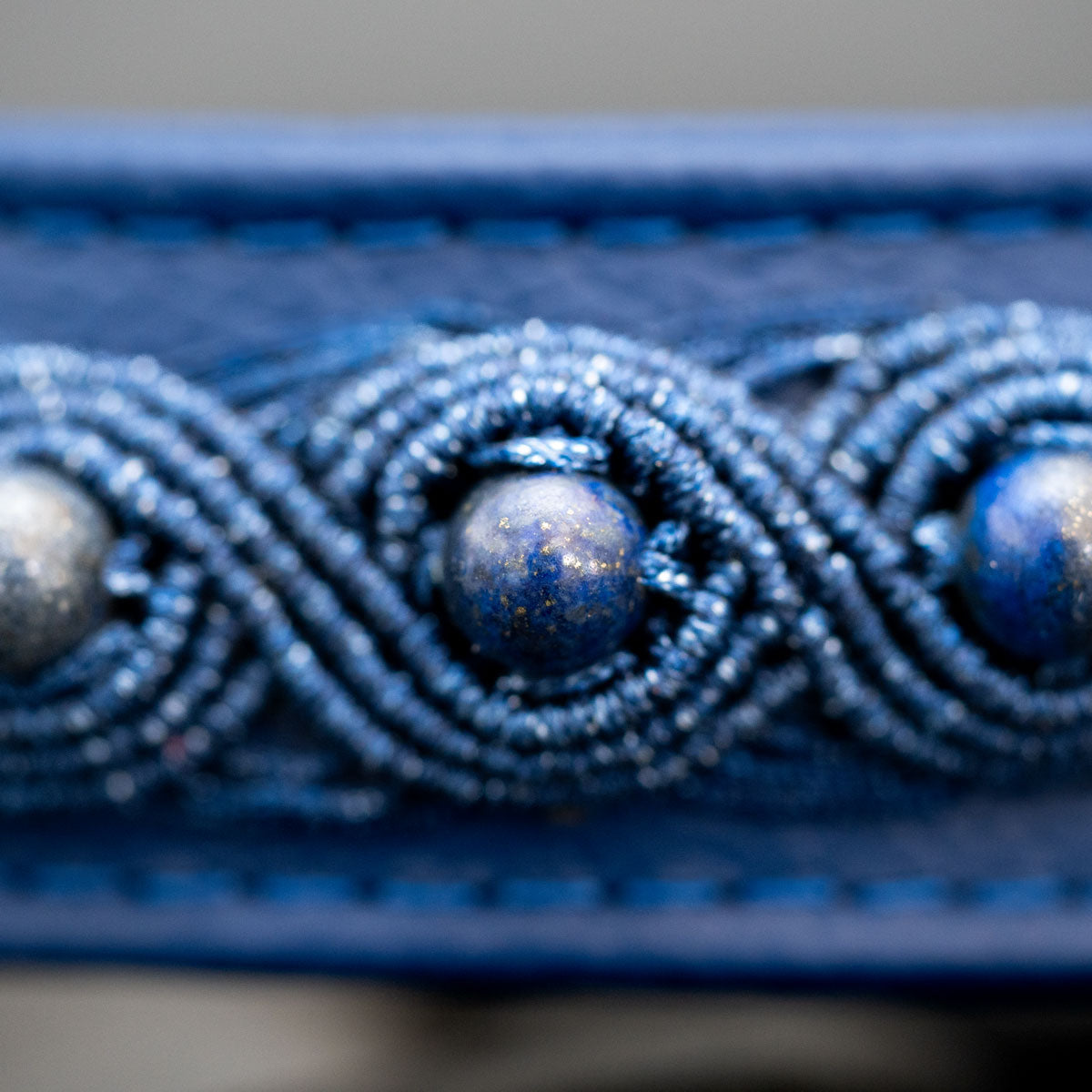 Makroaufnahme einer blauen Lapislazuli-Perle mit goldfarbenen Sprenkeln, eingefasst von schimmernder ultramarinblauer Micro-Makramee.