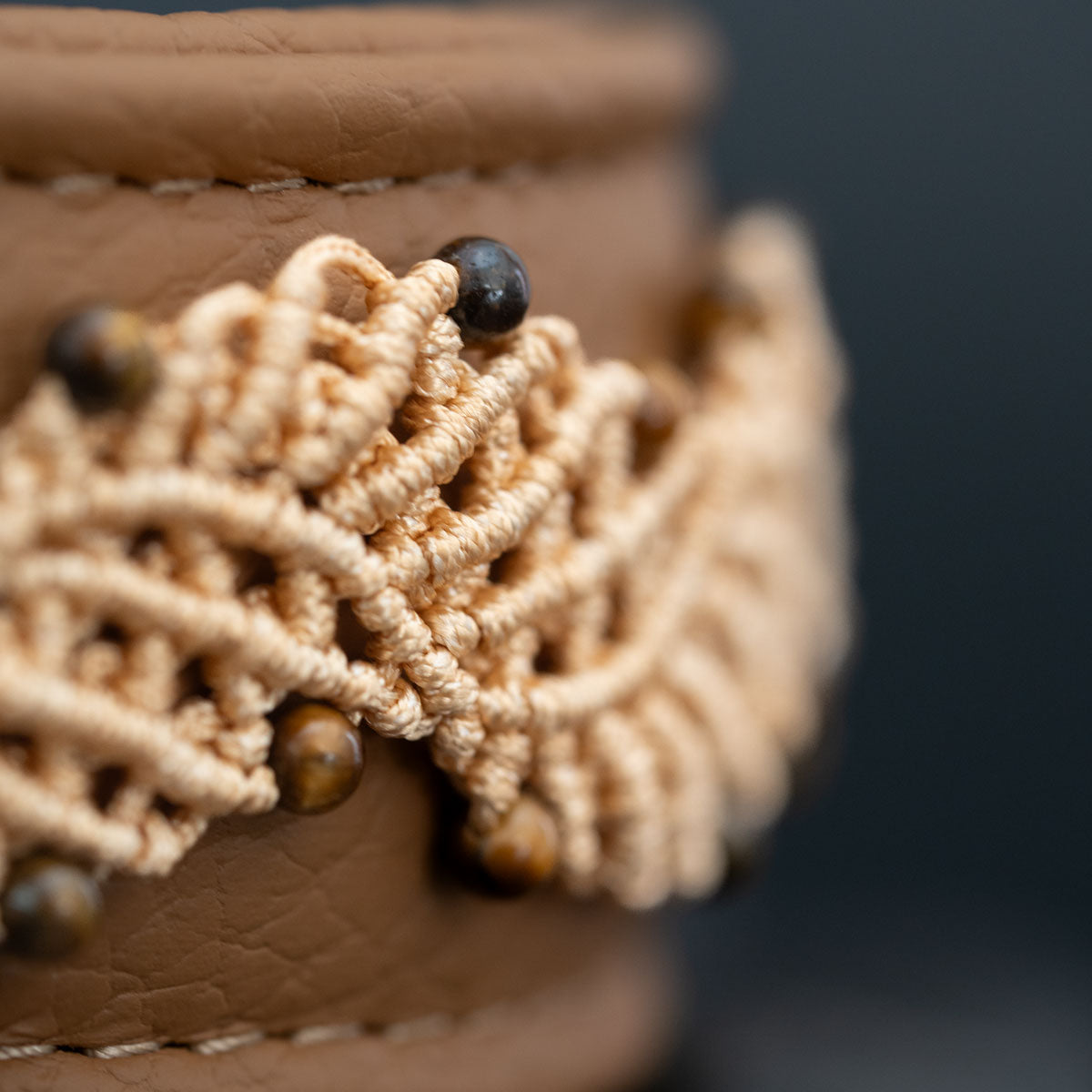 Seitliche Makroaufnahme der beigen Makramee-Knoten und braunen Tigerauge-Perlen auf cappuccinofarbenem Leder.