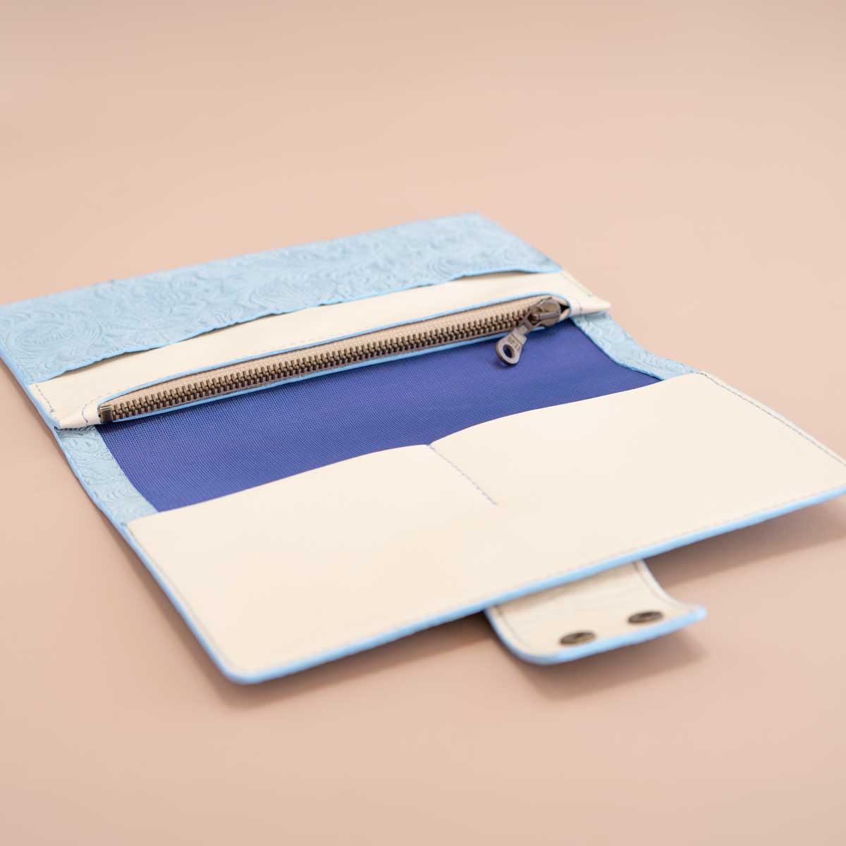 Geöffnetes, hellblaues Travel Wallet aus Leder. Die Innenansicht zeigt cremefarbenes Kalbsnappa, ein Reißverschlussfach mit blau-violett changierendem Futter und praktische Kartenfächer.