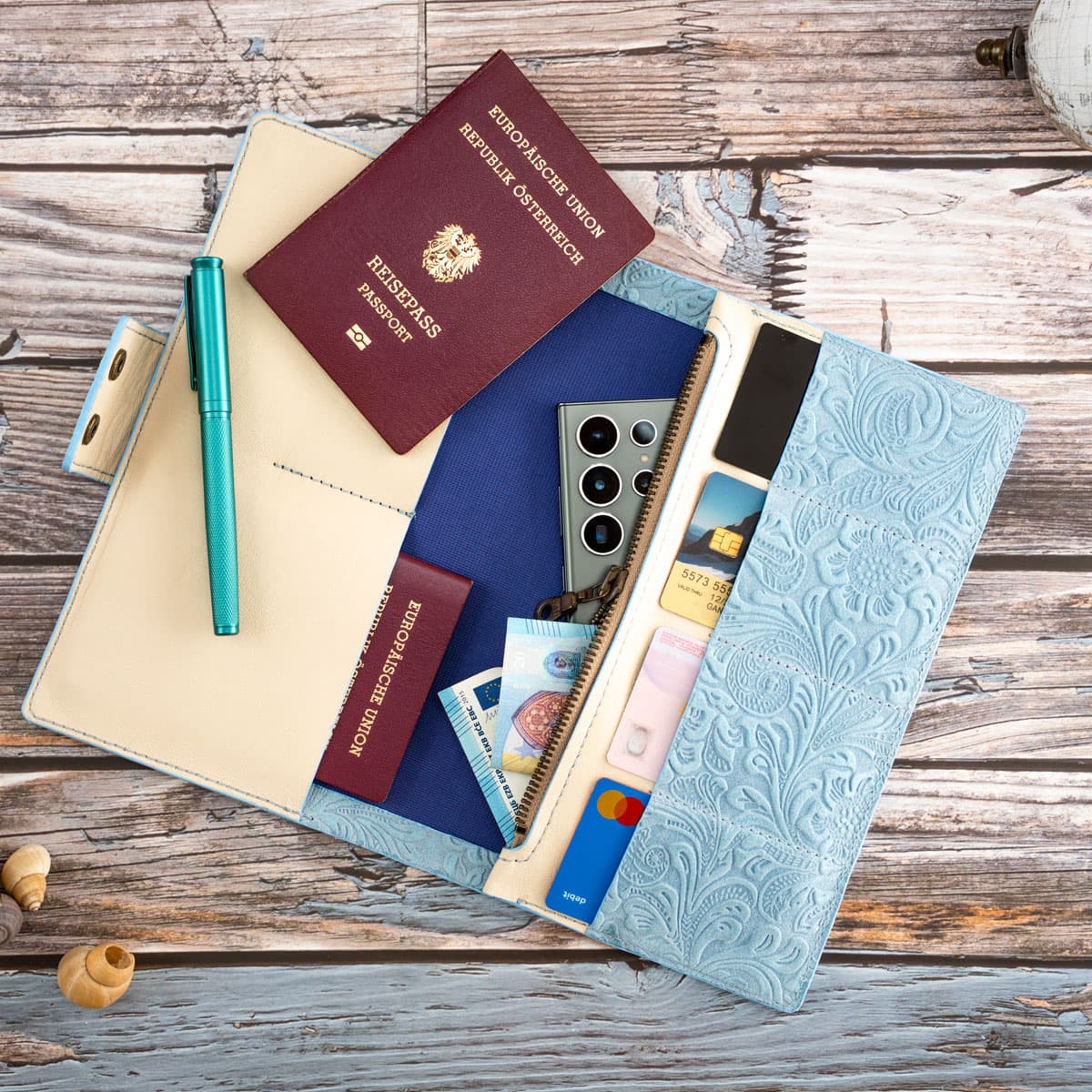 Befülltes Reise-Etui aus floral geprägtem Leder in Hellblau. Perfekt organisiert mit Platz für zwei Reisepässe, Kreditkarten, Bargeld und ein Smartphone.