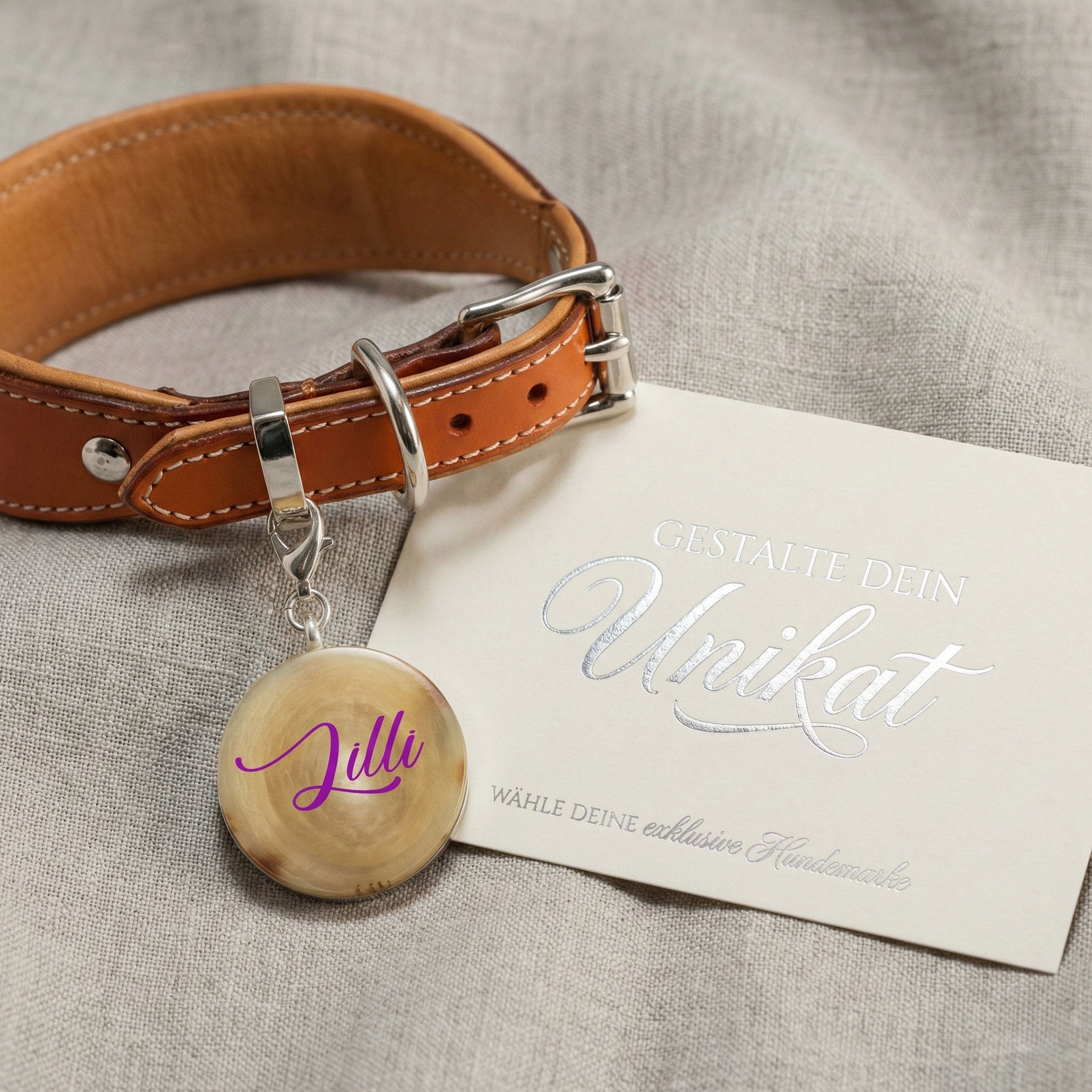 "Personalisiertes Set bestehend aus einer Büffelhorn-Hundemarke mit dem Namen 'Lilli' an einem braunen Lederhalsband mit silbernen Beschlägen. Daneben liegt eine edle Karte mit silberner Prägung 'Gestalte dein Unikat' auf grauem Leinenstoff."