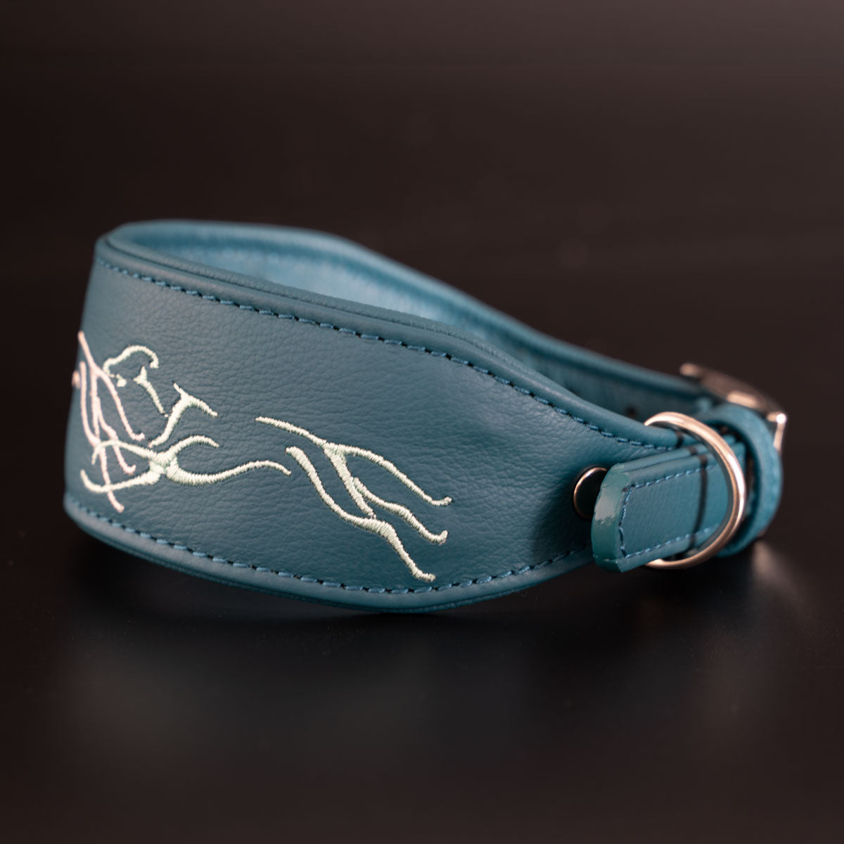 Seitliche Ansicht des petrolfarbenen Greyhoundline-Windhundhalsbands in geschwungener Fischform mit heller Greyhound-Stickerei und silberfarbenem Leinenring.