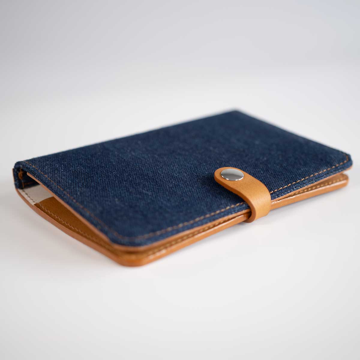 Vorderseite der geschlossenen Heimtierausweishülle aus robustem, dunkelblauem Denim-Stoff und cognacfarbenem Leder mit silbernem Druckknopf.