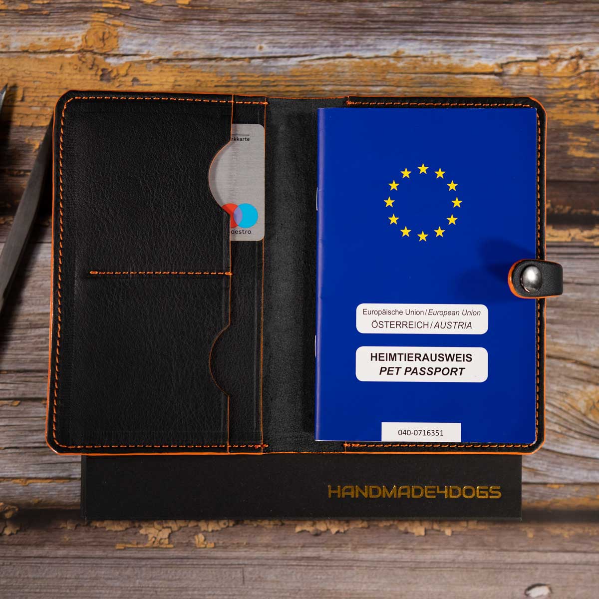 Geöffnete schwarze Heimtierausweishülle aus Leder mit orangefarbenen Kontrastnähten und einem sicher eingesteckten blauen EU-Heimtierausweis.