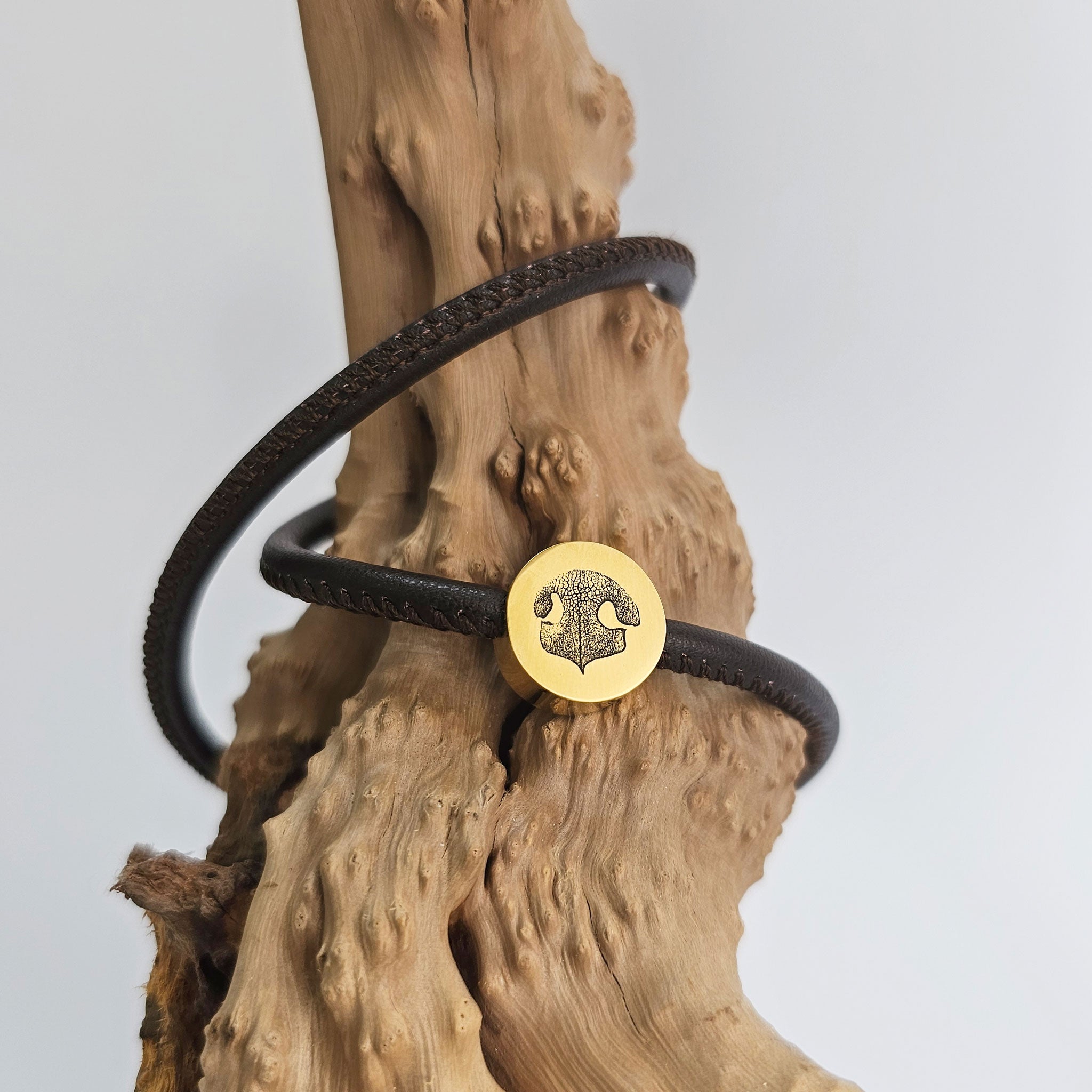 Dunkelbraunes Wickelarmband aus Leder mit goldener Hundenasen-Gravur, dekoriert auf einem Stück Treibholz