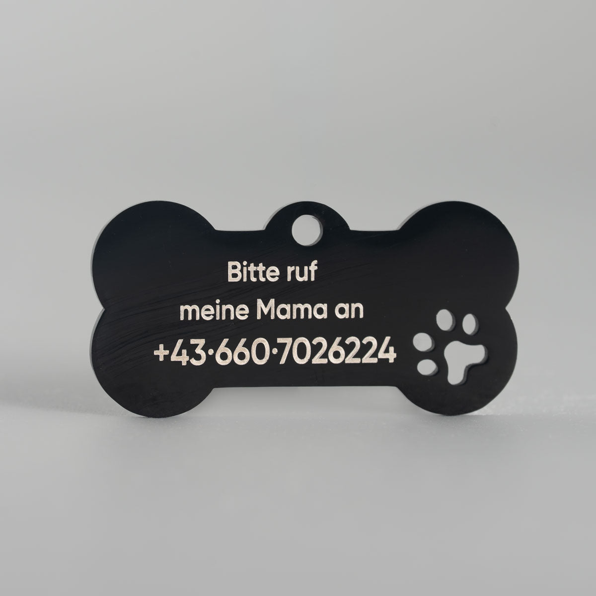 Schwarze Hundemarke aus Edelstahl mit hell gravierter Kontaktinformation und ausgestanzter Pfote auf der Rückseite.