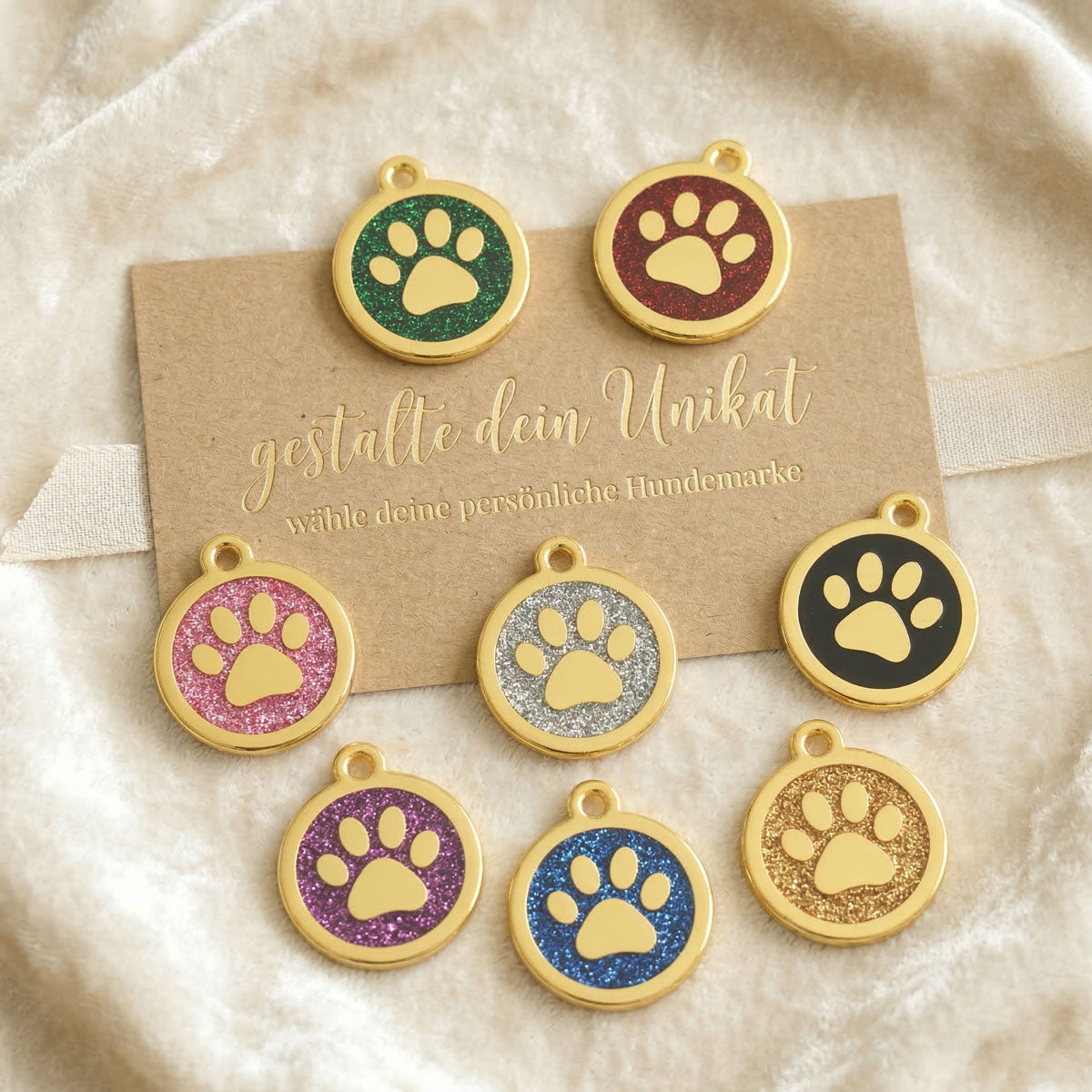Übersicht von acht personalisierbaren goldenen Hundemarken mit Pfoten-Motiv in verschiedenen Glitzer-Farben auf einer Präsentationskarte.