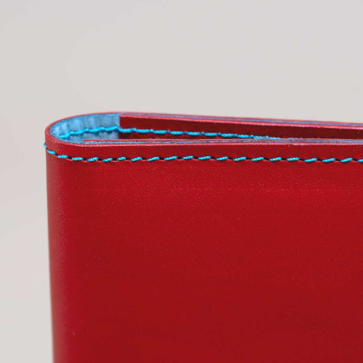 Detailansicht der präzisen hellblauen Kontrastnaht und des edlen silbernen Druckknopfs auf dem leuchtend roten italienischen Taschenleder.