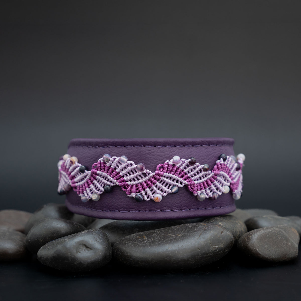 Amethyst-Halsband aus violettem Leder mit fliederfarbener und violetter Knüpfborte, auf Steinen vor dunklem Hintergrund.