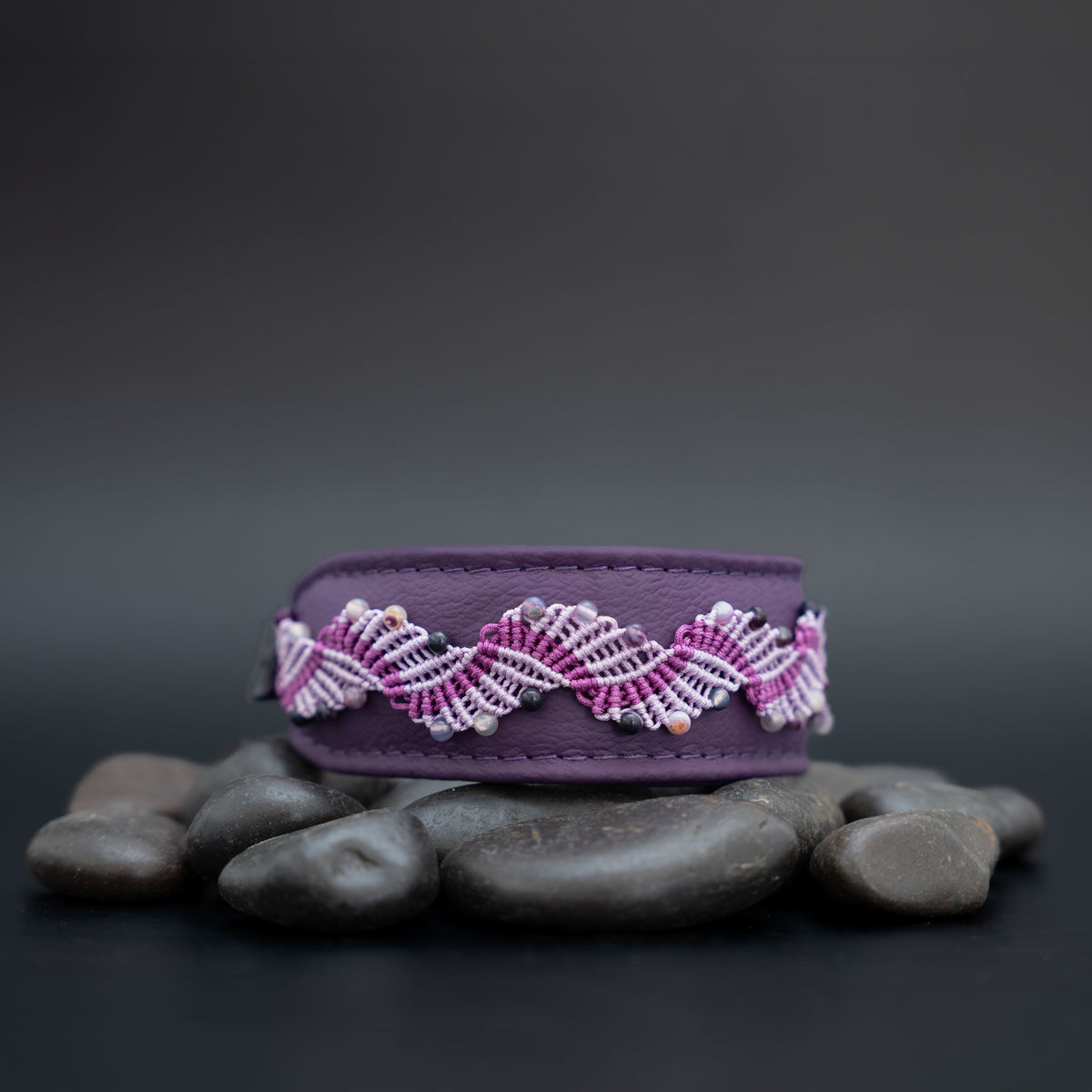 Amethyst-Halsband aus violettem Leder mit fliederfarbener und violetter Knüpfborte, auf Steinen vor dunklem Hintergrund.
