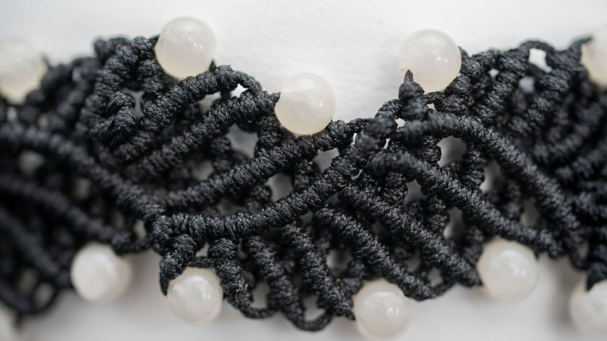 Detail im Querformat: geschwungene schwarze Knotenreihen und helle Mondsteine am weißen Lederhalsband.