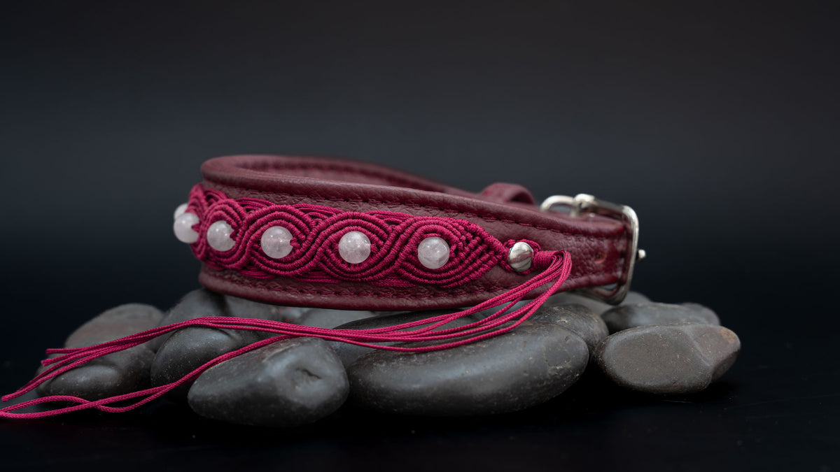Rubinrotes Makramee-Hundehalsband mit Rosenquarz im Querformat; lange Garnenden liegen vor dem Halsband auf dunklen Steinen.