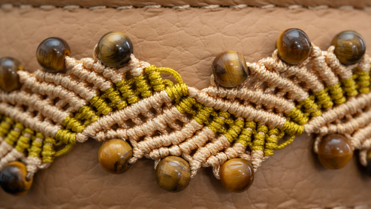 Detail im Querformat: blattartige Makramee-Motive in Eierschale und Gold mit Tigerauge-Perlen vor cappuccinofarbenem Leder.
