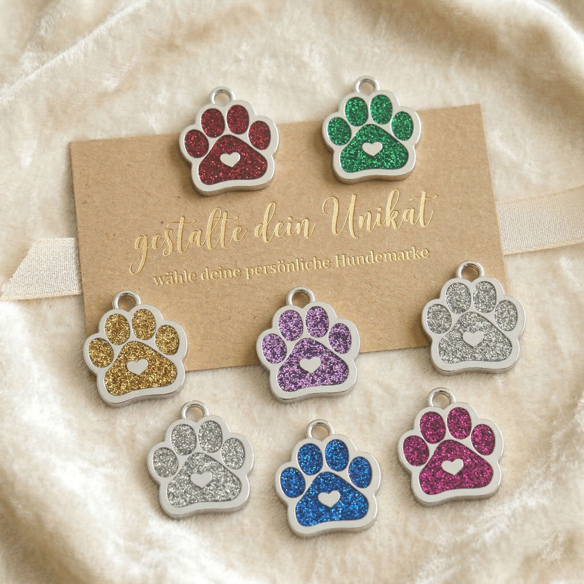 Sortiment von glitzernden Hundemarken in Pfotenform in den Farben Rot, Grün, Gold, Silber, Blau und Magenta auf einer Karte mit der Aufschrift „gestalte dein Unikat“.