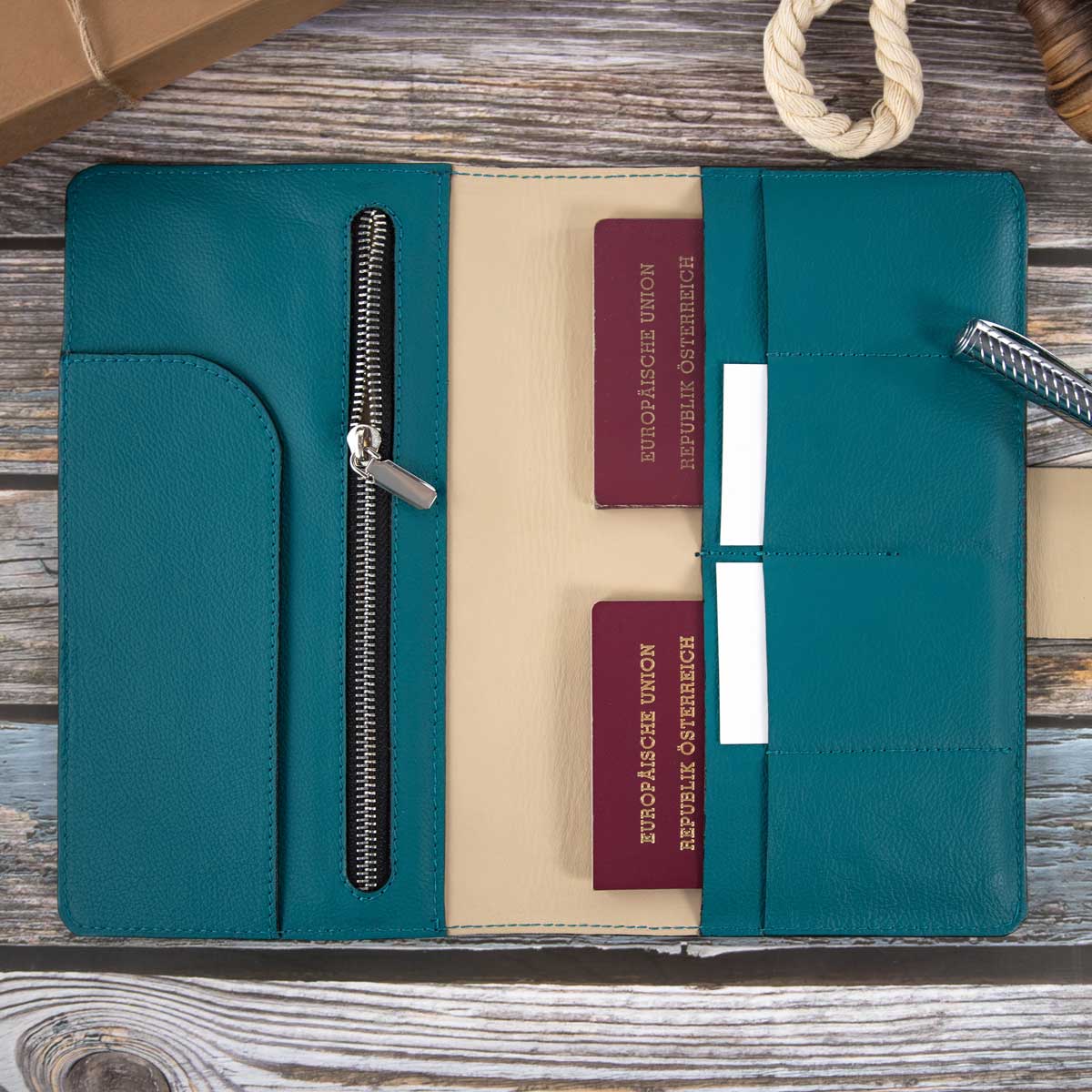 Geöffnetes Leder Travel Wallet in Petrol und Taupe, befüllt mit zwei österreichischen Reisepässen und einem Stift, liegend auf einem rustikalen Holztisch