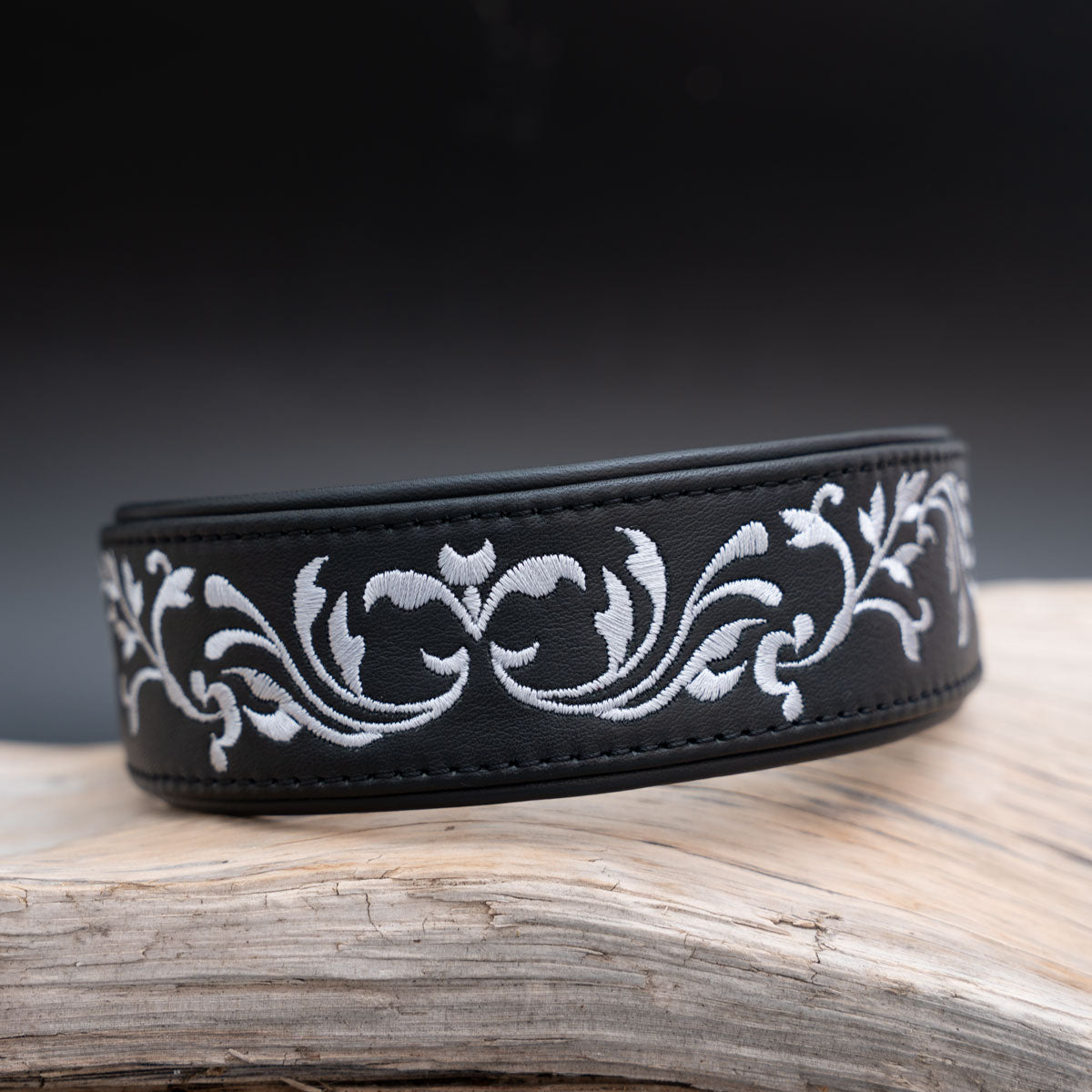 Schwarzes Windhundhalsband mit silberner Ornamentstickerei, schräg von vorne auf hellem Holz fotografiert.